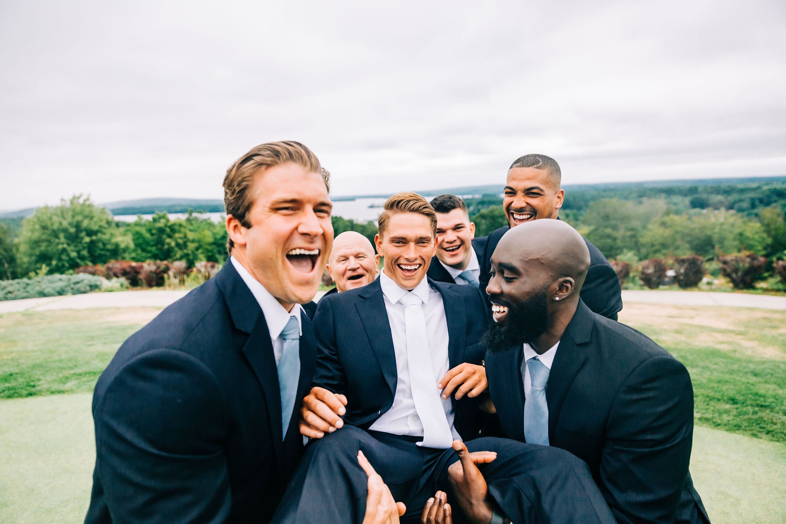 Joyful groom lifted by laughing groomsmen in navy suits during outdoor wedding celebration with scenic backdrop.