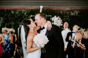 Bride and groom sharing first kiss holding pink bouquet while guests celebrate at wedding reception.