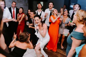 Bride in white jumpsuit singing with microphone on dance floor surrounded by wedding guests.