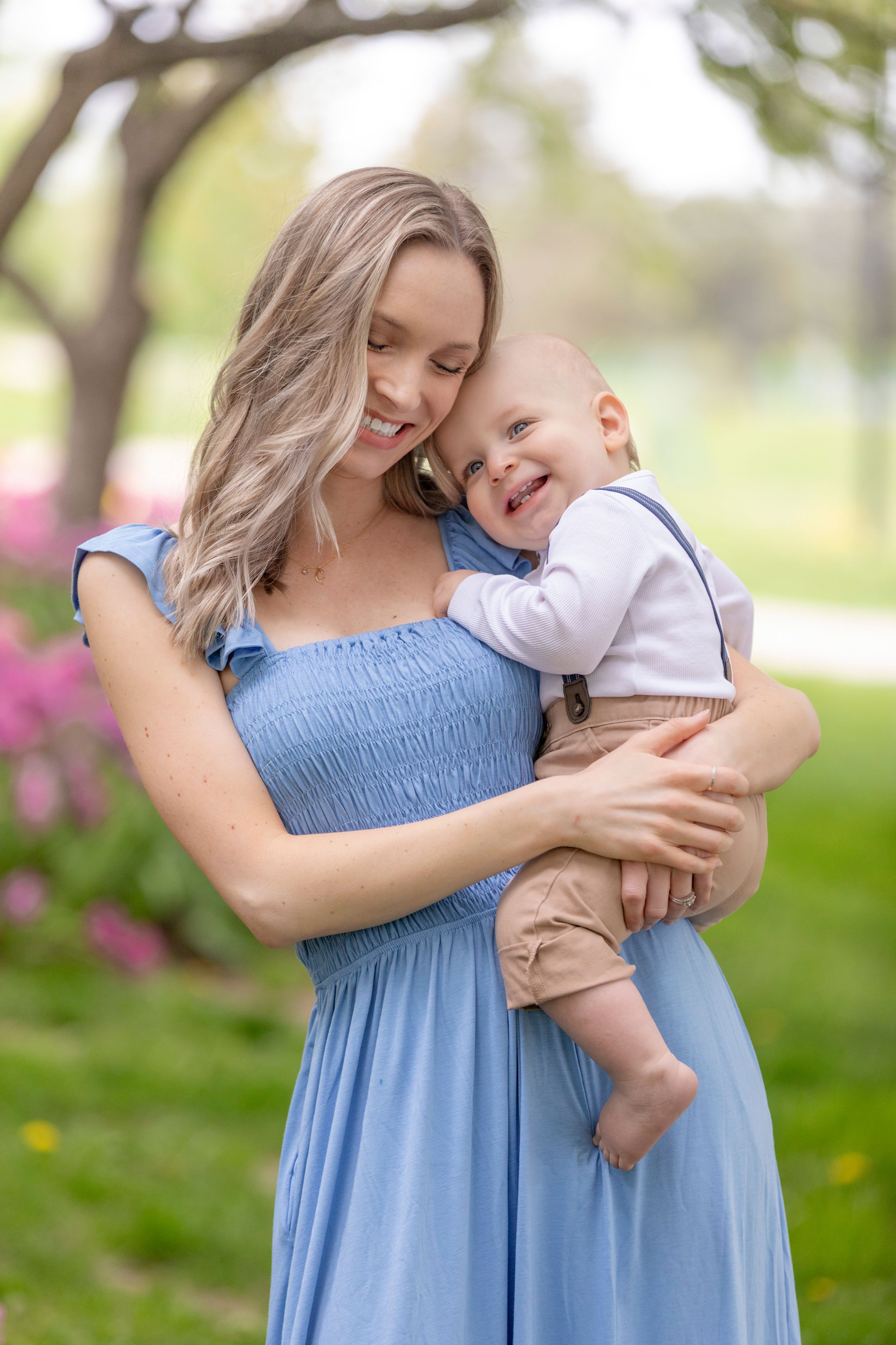 Smiling blonde mother in blue dress holding happy baby boy in a blooming spring park portrait.