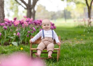 Adorable baby boy in suspenders sits on a wooden bench surrounded by purple tulips in a park.