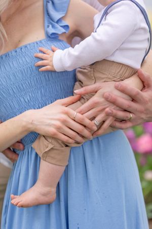 Parents holding a baby together, mother in blue dress with wedding rings visible on their hands.