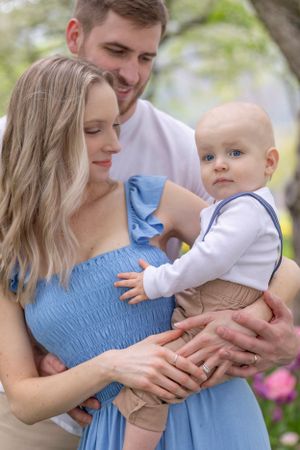 Young family with blue-eyed baby boy in suspenders, mother in blue dress, outdoors in spring.
