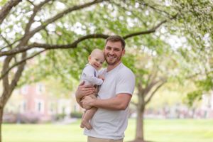 Smiling father holding infant baby while standing under blooming trees in a green park.