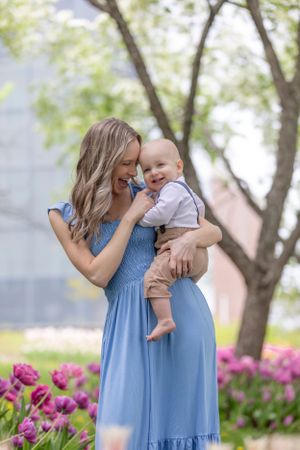 Smiling mother in blue dress holds laughing baby boy among pink tulips in a garden setting.