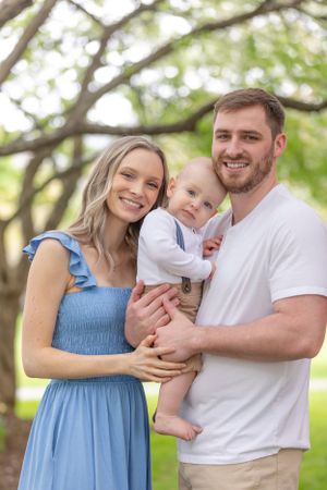 Smiling young family with mother in blue dress and father in white tee holding baby outdoors.
