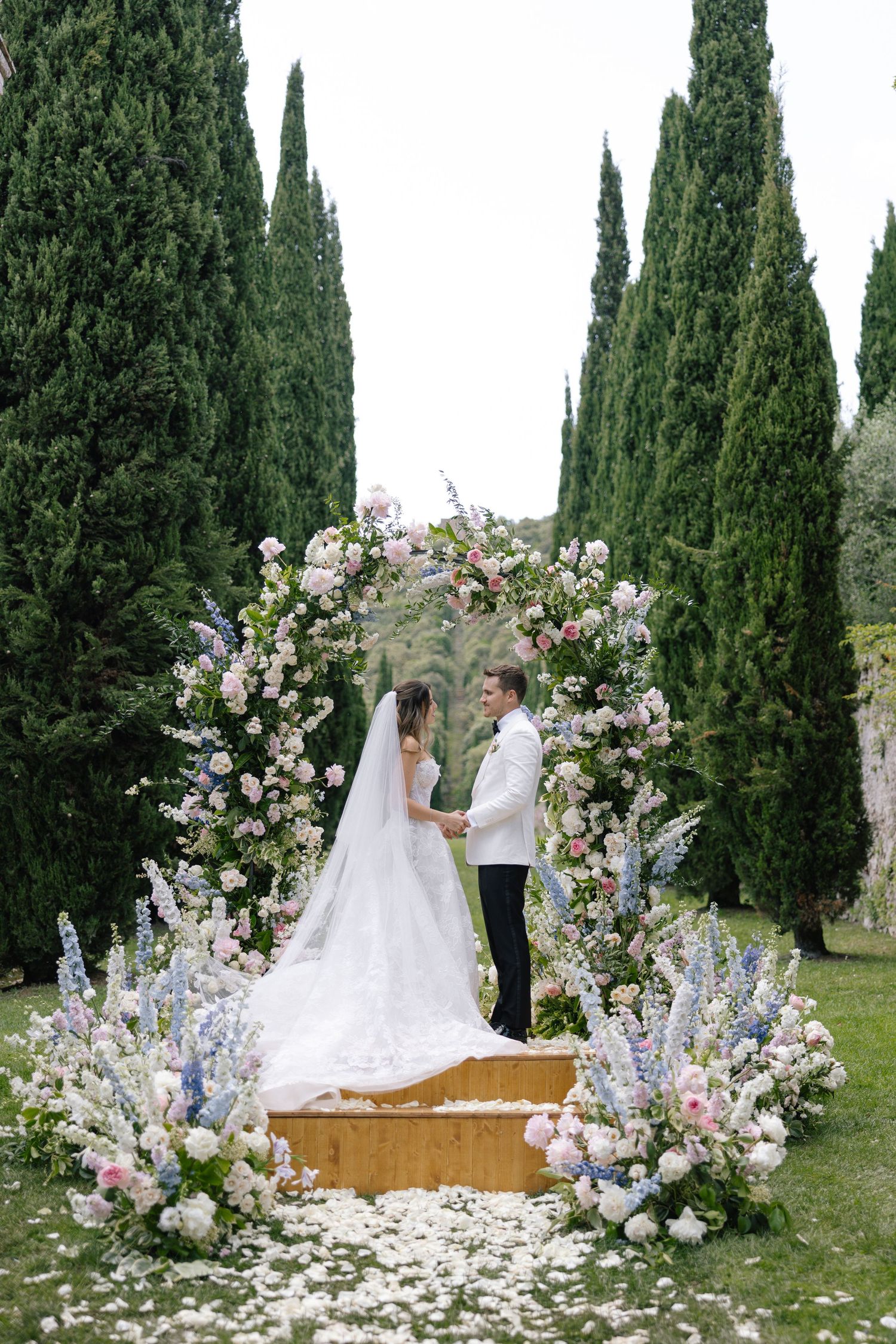A couple walks down an outdoor aisle lined with flowers and cypress trees leading to an elegant villa entrance.