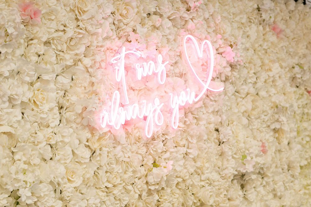 Pink neon sign reading 'it was always you' on a white floral wall backdrop.