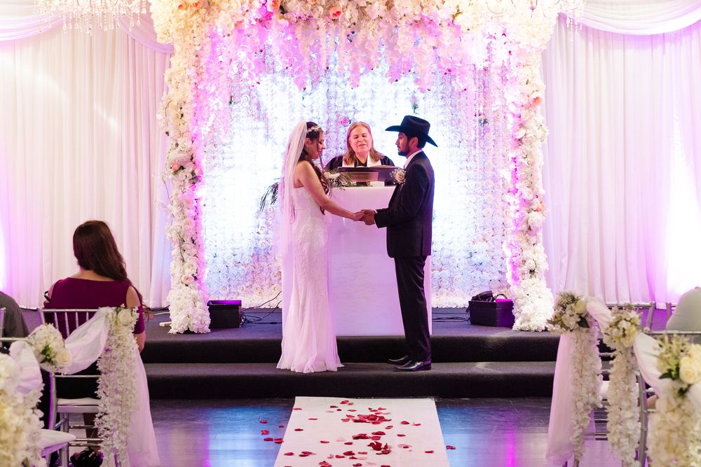 Bride and groom exchange vows at floral wedding altar with pink roses and white flowers under purple lighting.