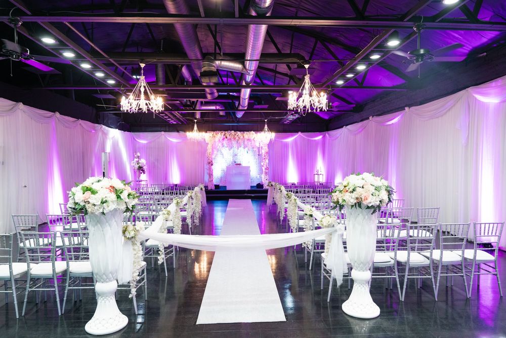 Elegant wedding venue with purple lighting, white floral arrangements, chiavari chairs, and a white aisle runner leading to a draped altar.