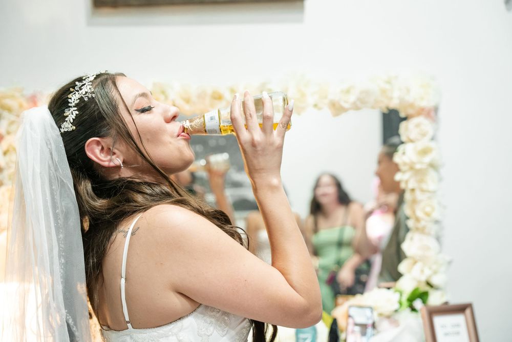 Bride in white gown and veil drinking from a bottle during wedding preparations near a floral mirror.