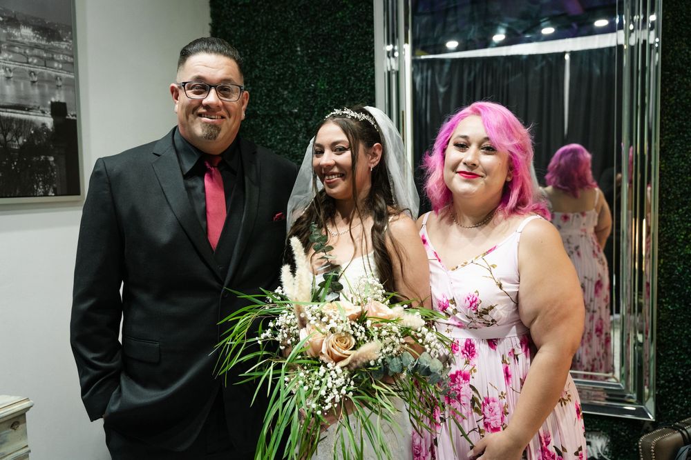 Bride in white gown with bouquet poses with man in black suit and woman in pink floral dress at wedding venue.