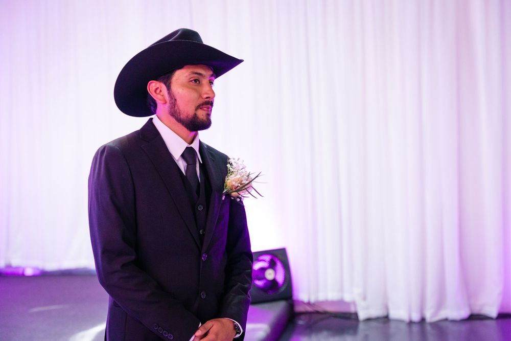 Man in black suit and cowboy hat with boutonniere stands at a wedding ceremony.