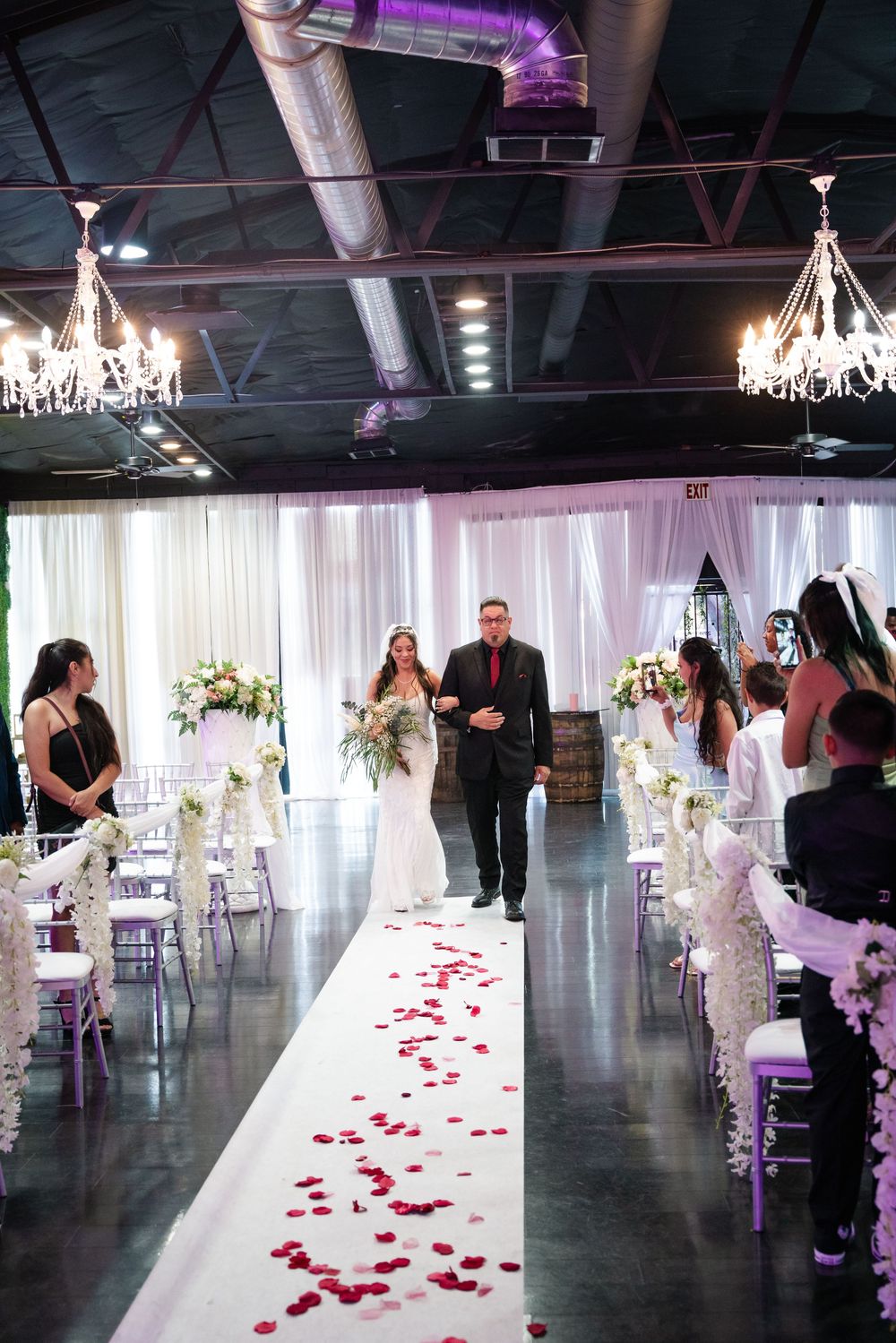 Bride walks down rose petal aisle with escort in elegant indoor wedding venue with chandeliers and white draping.