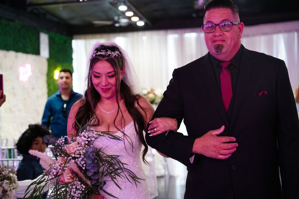 Bride in white gown walks down aisle with escort in black suit at elegant wedding reception venue.