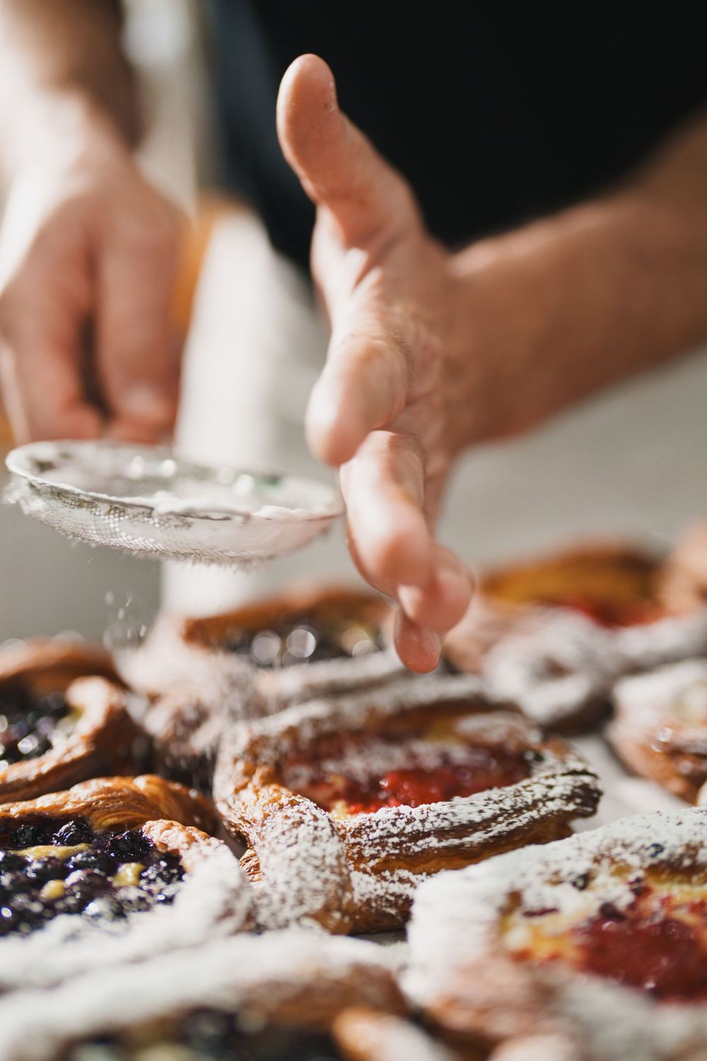Baker dusting powdered sugar over fresh fruit pastries using a fine mesh sieve.