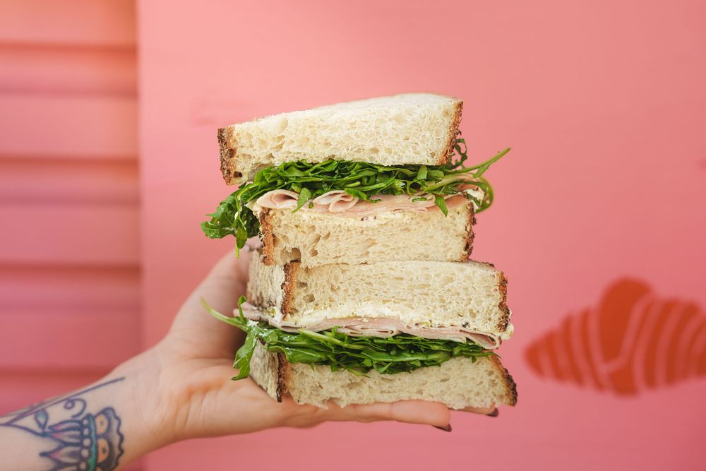 Tattooed hand holding a stacked sandwich with arugula on whole grain bread against a pink wall.
