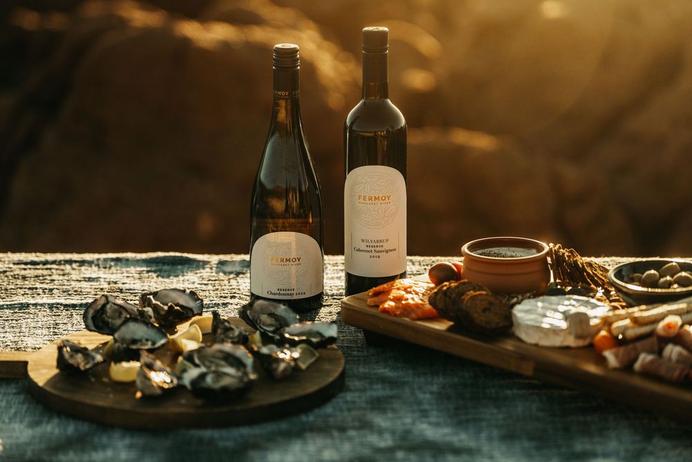 Two wine bottles beside oysters and charcuterie board in golden sunset light.