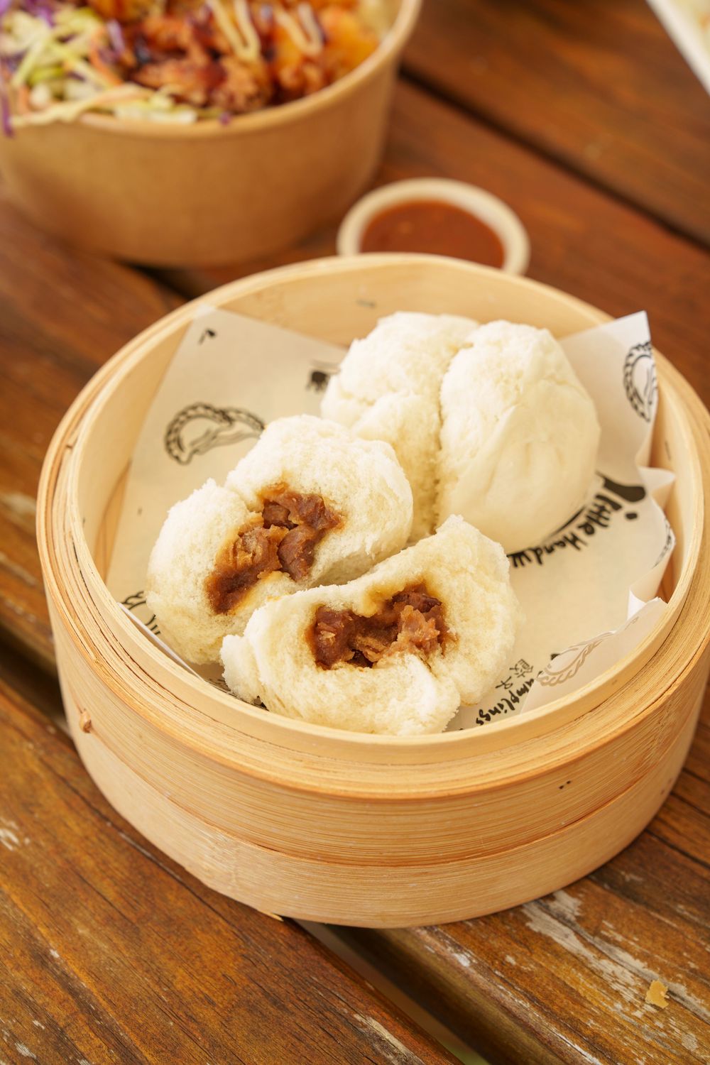 Steamed pork bao buns in a bamboo steamer basket showing filling on wooden table.