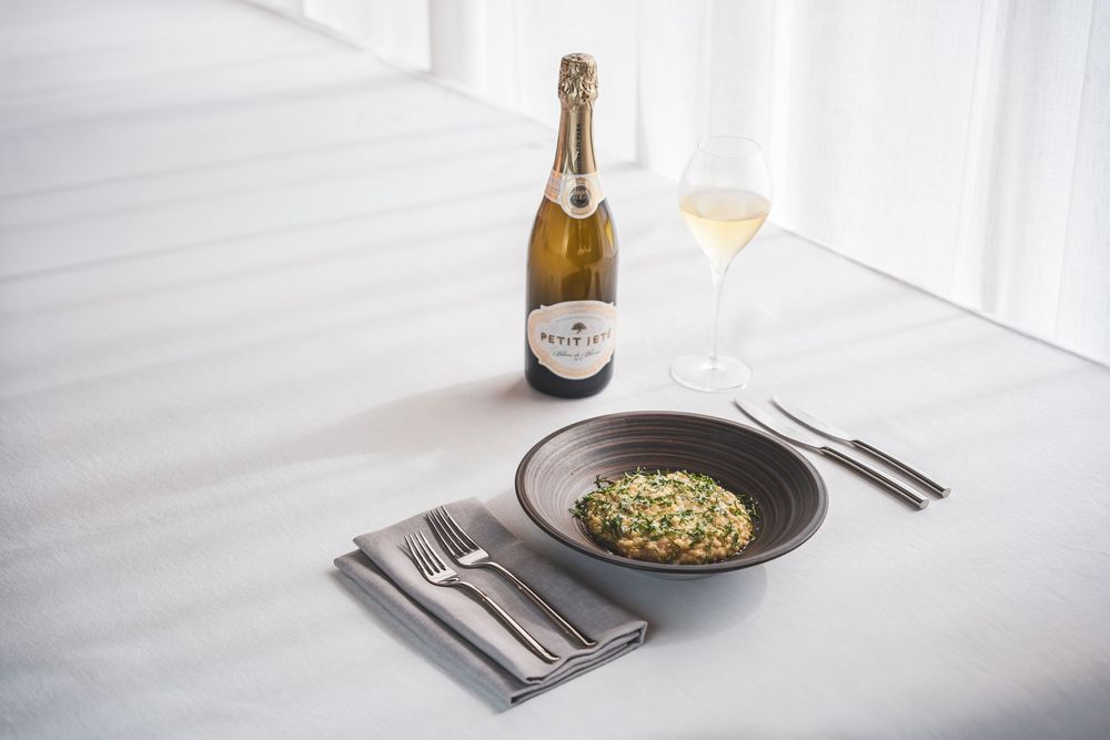 Green herb risotto in dark bowl paired with Petit Jean sparkling wine on white table.