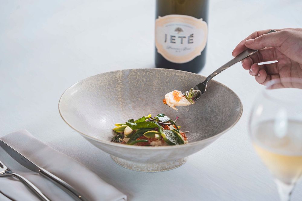 Hand spooning sauce over elegant seafood dish paired with Jete white wine bottle.