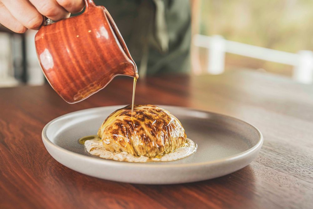 Chef pouring brown butter sauce from a red ceramic jug over a roasted cauliflower dish on a plate.