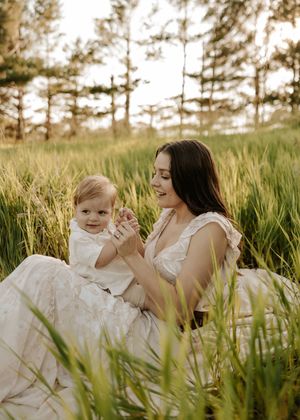A young mother sits in a lush green field holding her baby, sharing a tender moment surrounded by tall grass and trees.