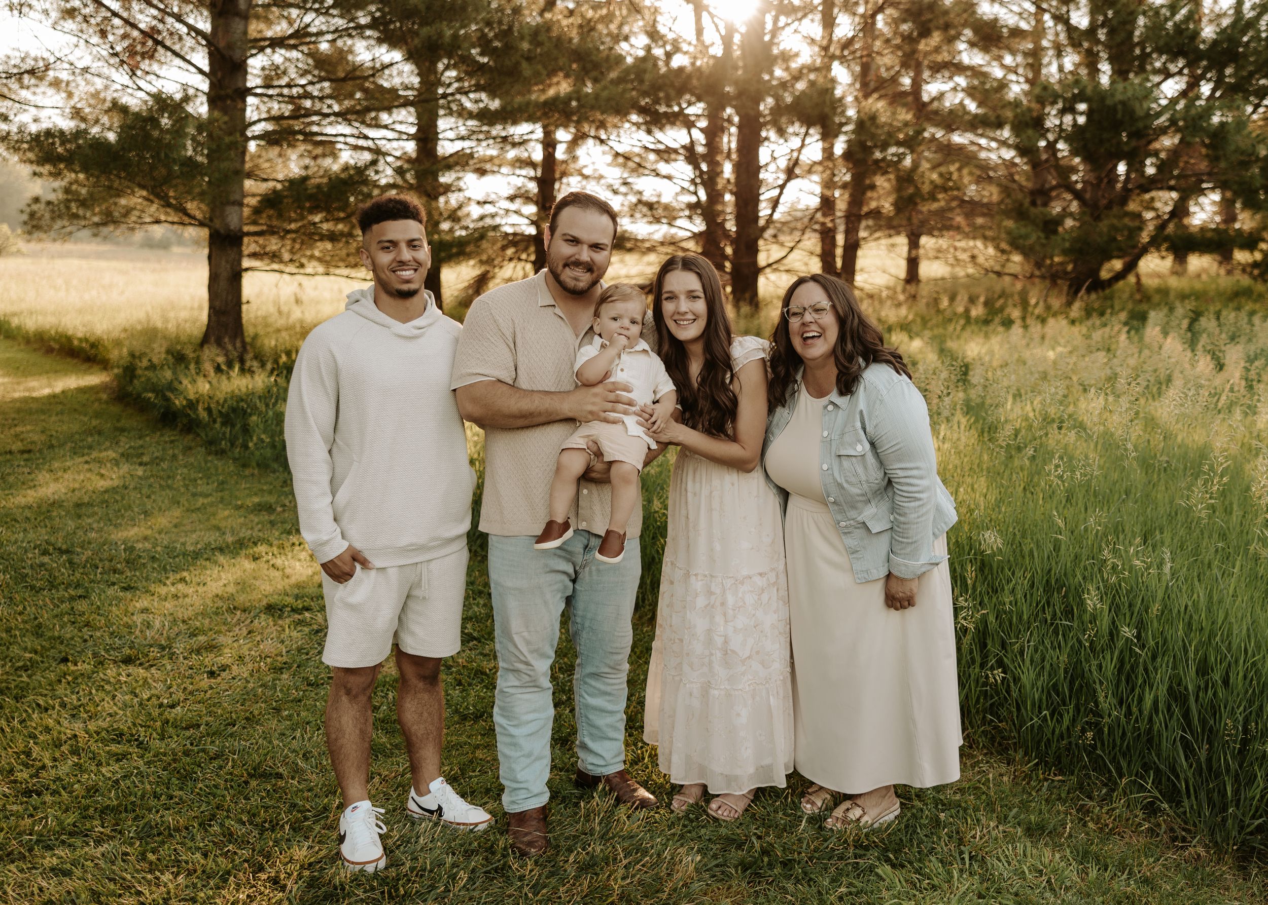 Family of five posing outdoors in a sunny field, wearing neutral tones, smiling together during golden hour.