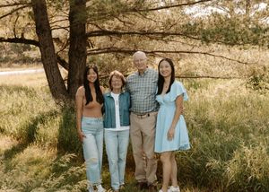 Family of four smiling outdoors under a pine tree in a sunny meadow, wearing casual summer outfits.