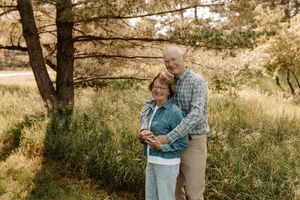 Elderly couple embracing outdoors near pine trees in a lush green meadow, smiling warmly in casual attire.