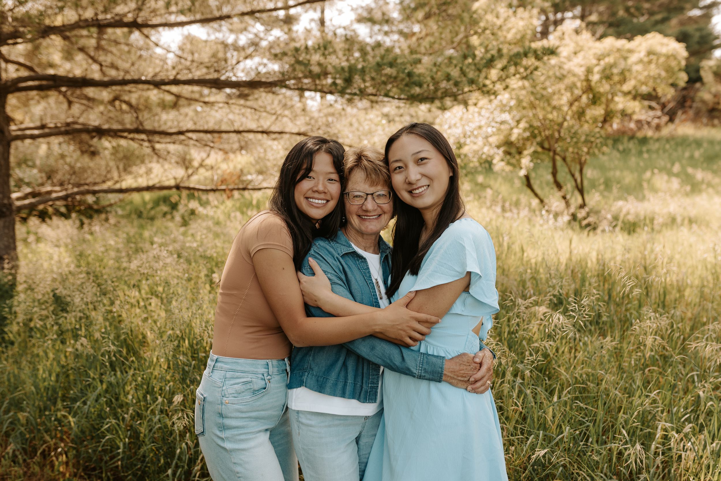 Three women share a warm embrace in a sunlit meadow surrounded by golden trees.