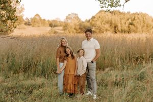 Family of four posing in a golden meadow at sunset, wearing warm earth tones, surrounded by tall grass and trees.