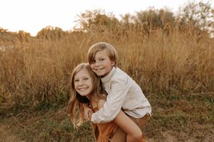Two smiling children share a piggyback ride in a golden field during sunset, wearing warm earth-toned outfits.