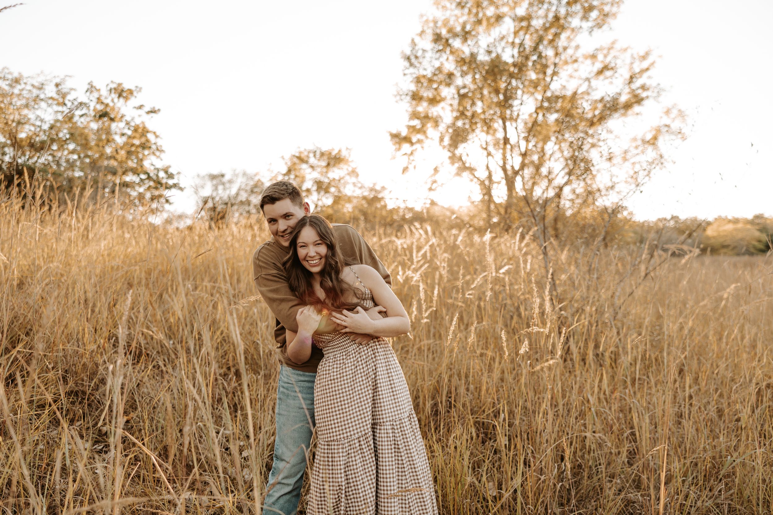 Couple embracing in golden field at sunset, woman in flowy dress, man in casual attire, surrounded by tall dry grass and trees.