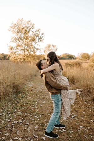 Couple sharing a romantic kiss in an autumn field, man lifting woman in a warm golden sunset outdoor setting.