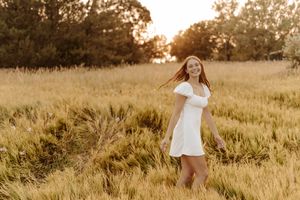 Young woman in white dress walking through golden field at sunset with trees in background.