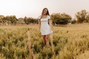 Young woman in white mini dress standing in golden field with wildflowers and trees at sunset.