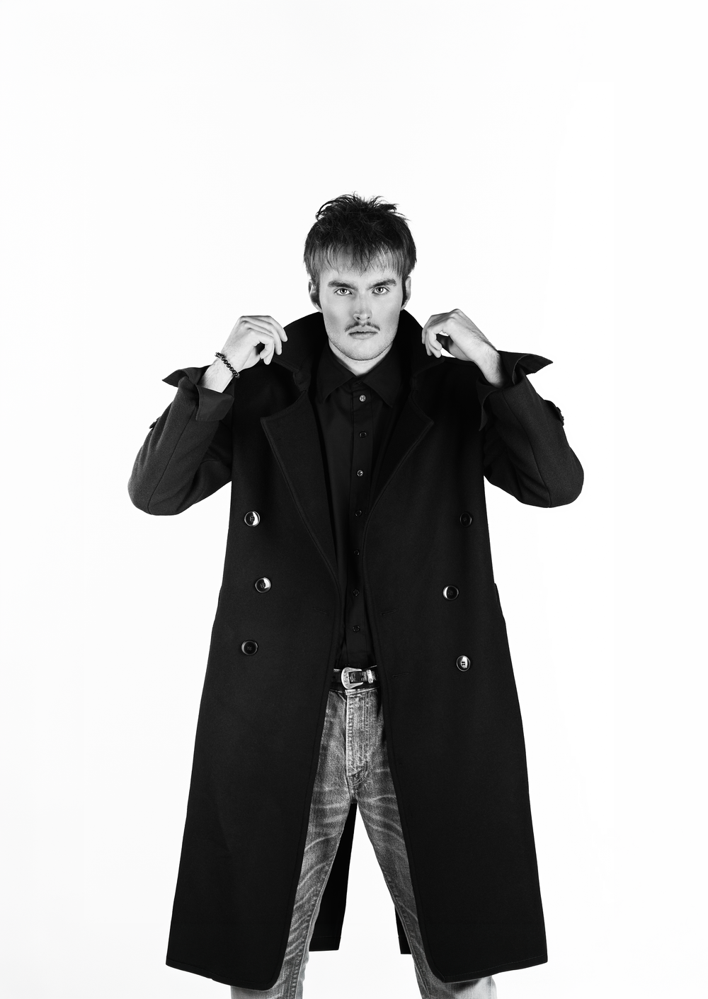 Man in long black double-breasted coat adjusting collar, wearing plaid trousers, black and white portrait photo.