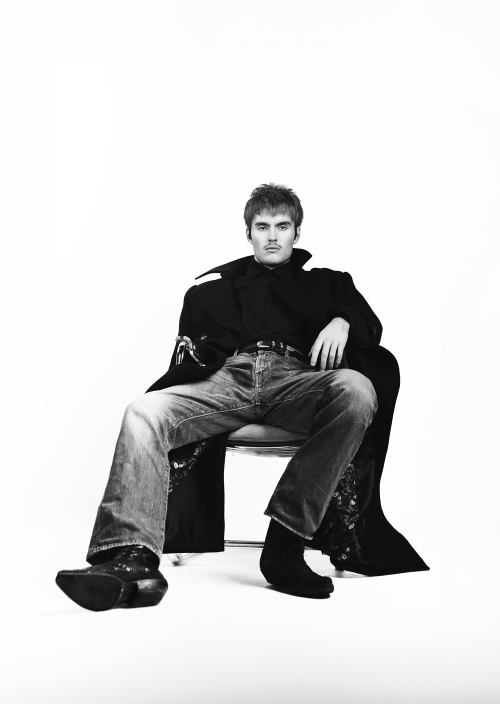 Young man in black coat and flared jeans sits confidently on a stool in a black and white studio photo.