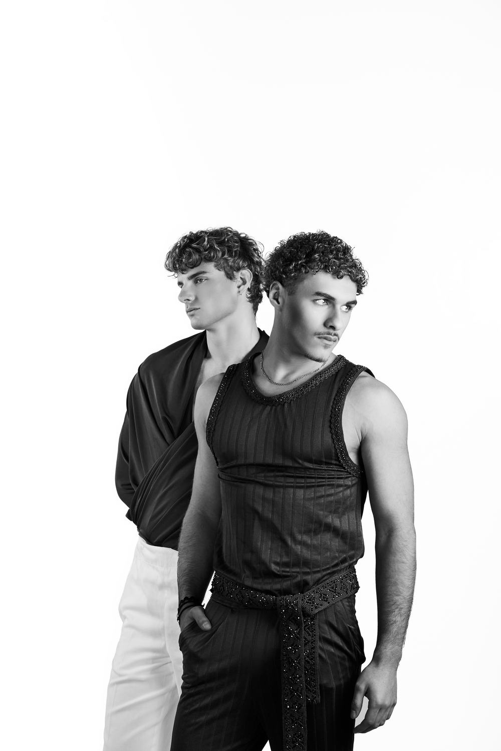 Two curly-haired men in stylish black outfits pose together in a black and white fashion portrait.