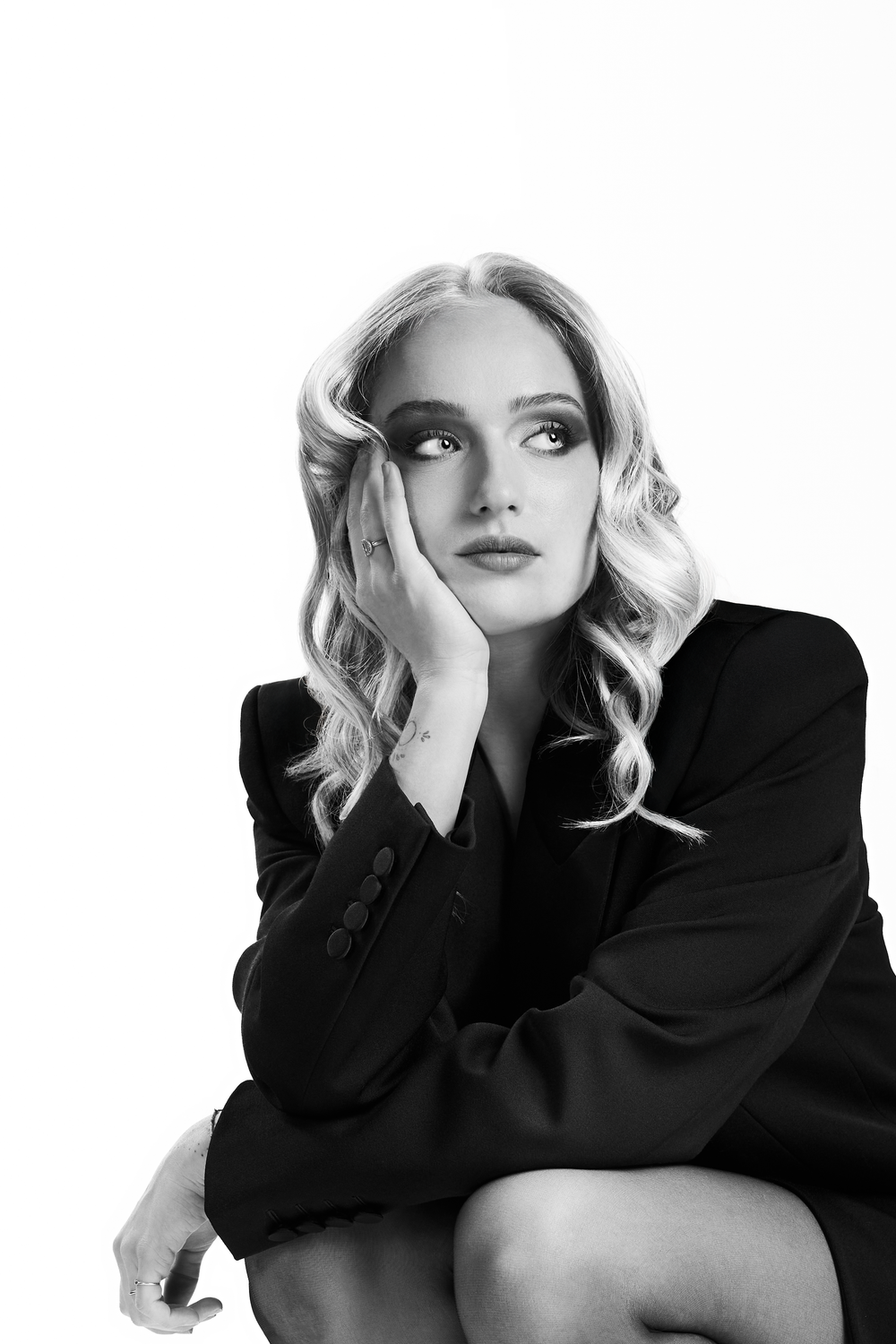 Black and white portrait of a blonde woman in a dark blazer, resting her cheek on her hand, gazing upward thoughtfully.