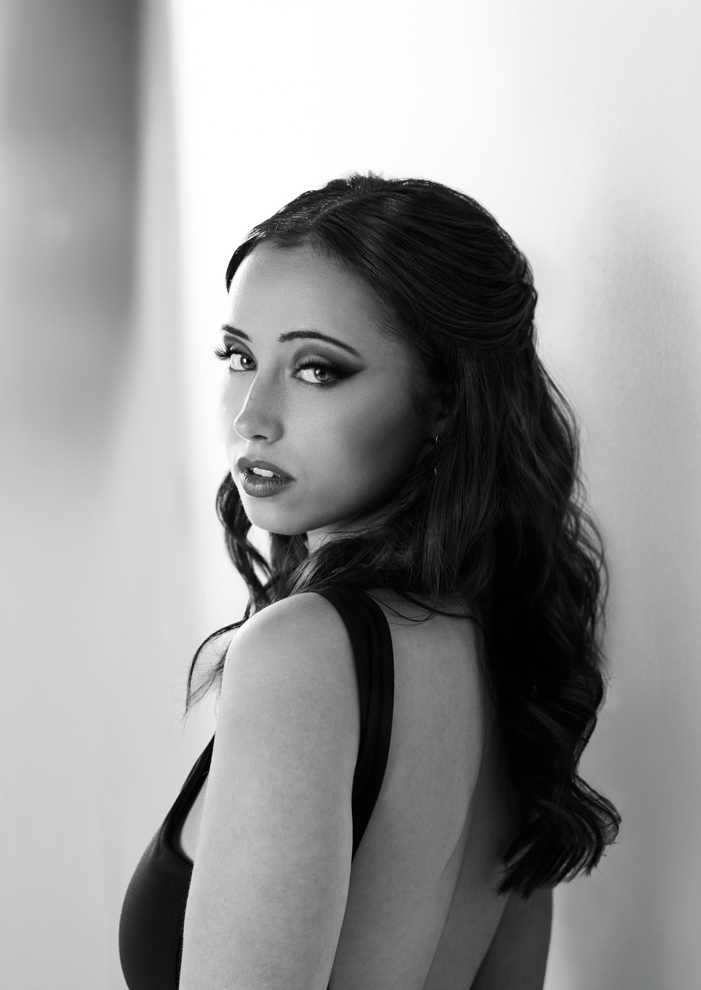 Black and white portrait of a young woman with long wavy dark hair, wearing a backless dress, looking over her shoulder.