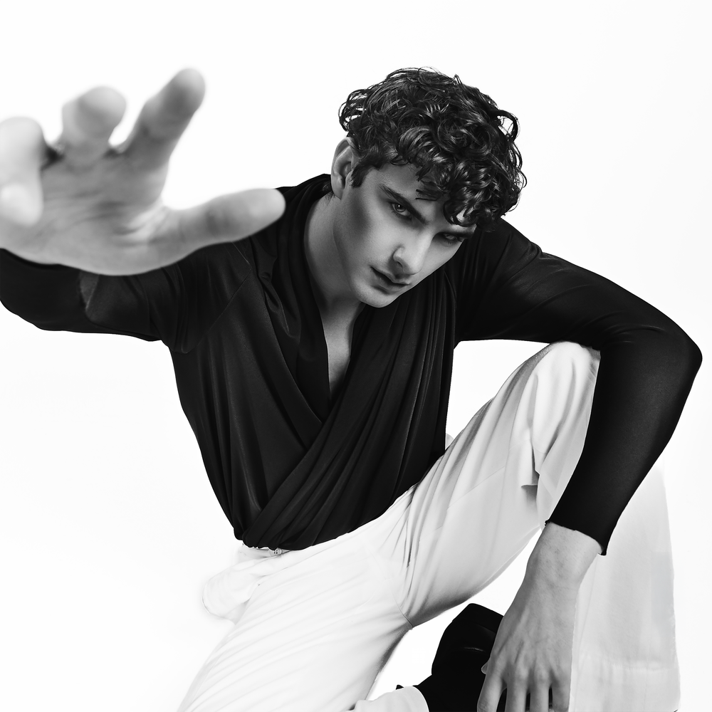 Young male dancer in black top and white pants reaches toward camera in dramatic black and white pose.
