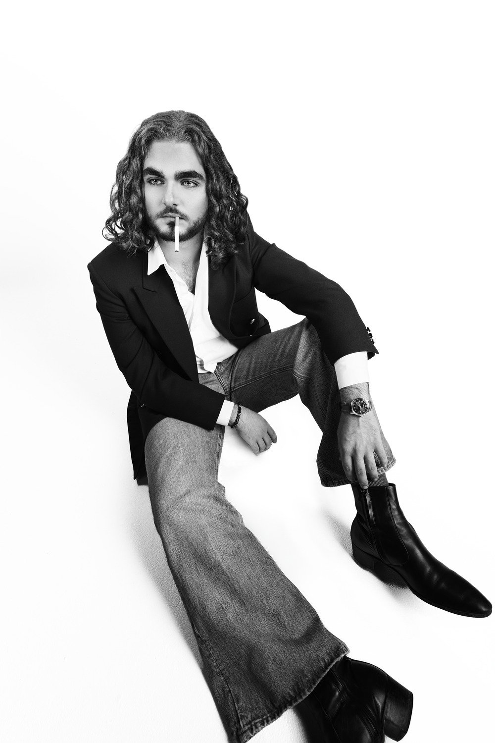 Black and white photo of a long-haired man in blazer and flare jeans sitting on floor with cigarette.