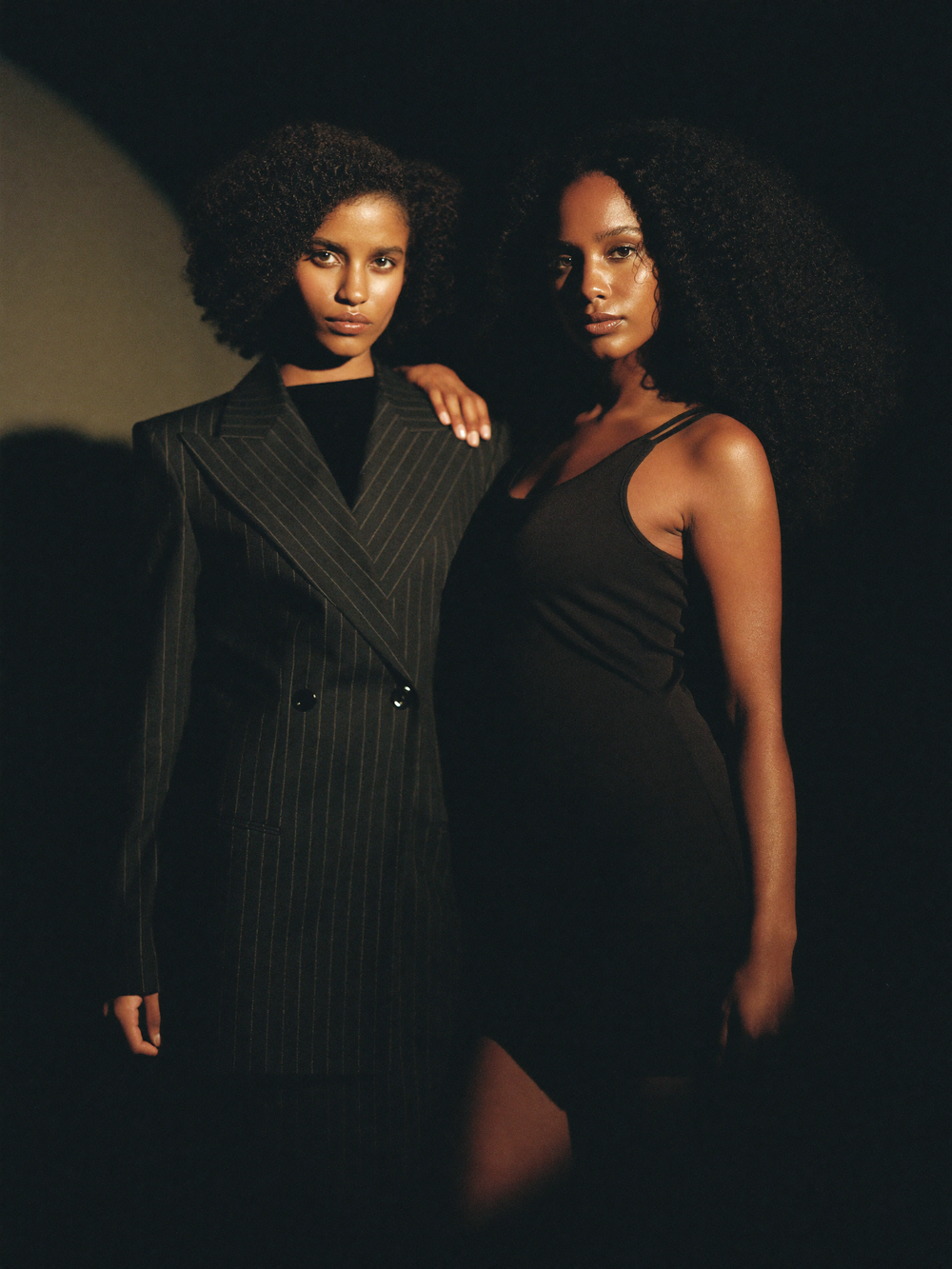 Two Black female models pose in dark outfits, one in pinstripe blazer dress, one in black mini dress, against dark background.