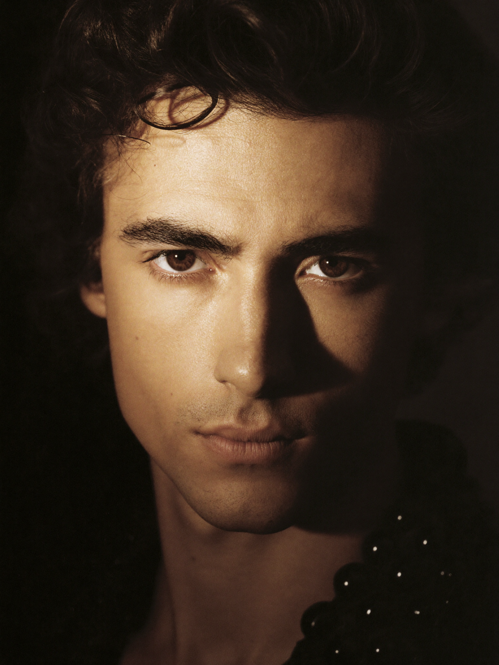 Close-up portrait of a young man with dark curly hair and brown eyes in dramatic low-key lighting.