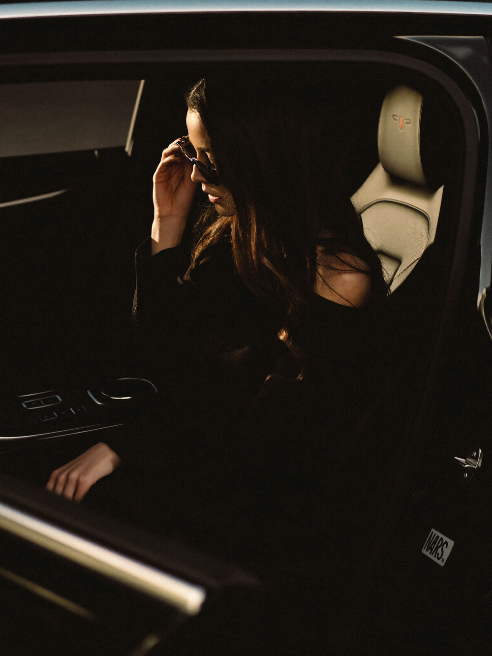 Woman in black outfit adjusting sunglasses while seated in luxury car with cream interior.