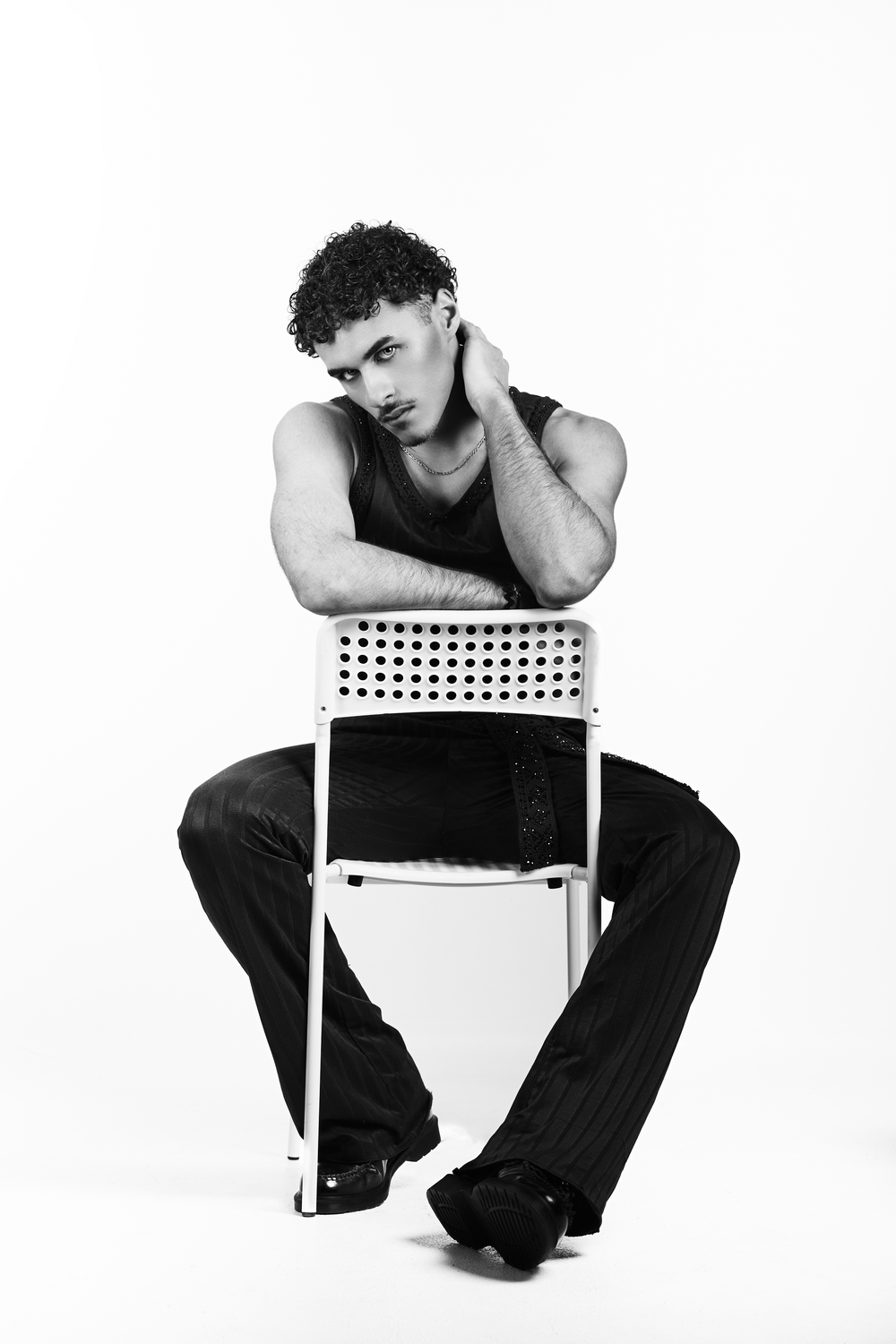 Black and white photo of a young man with curly hair sitting backwards on a chair in a sleeveless top and black pants.