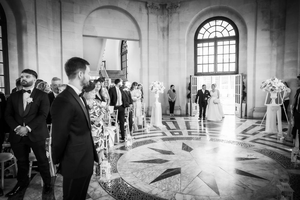 Black and white wedding ceremony photo in elegant hall with ornate floors, guests watching bride walk down the aisle.