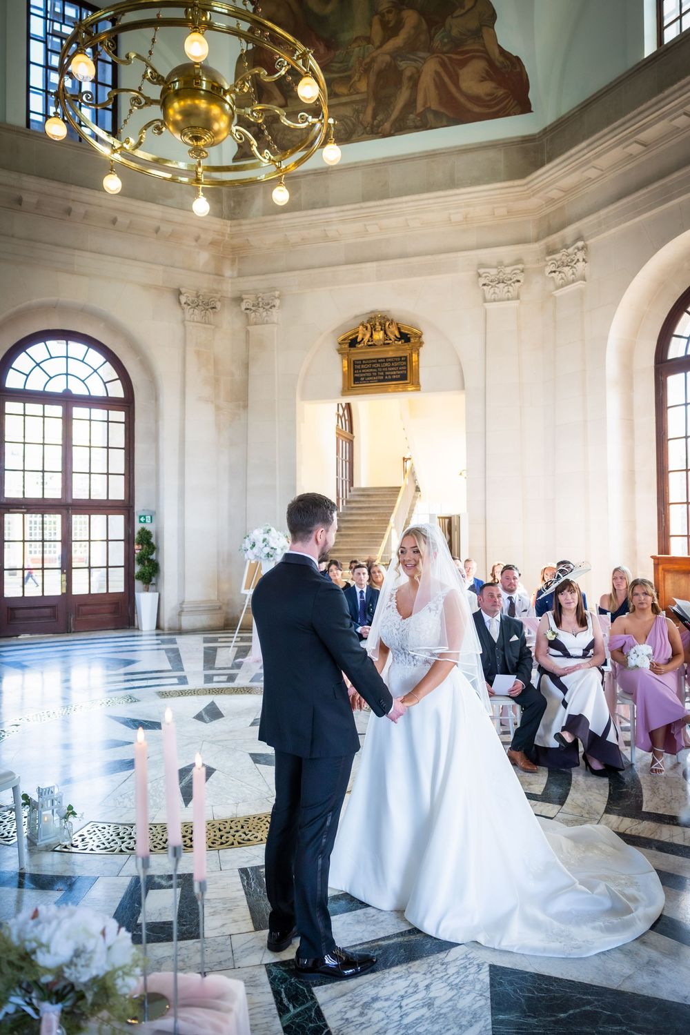 Bride and groom holding hands during elegant wedding ceremony in ornate marble hall with guests seated.